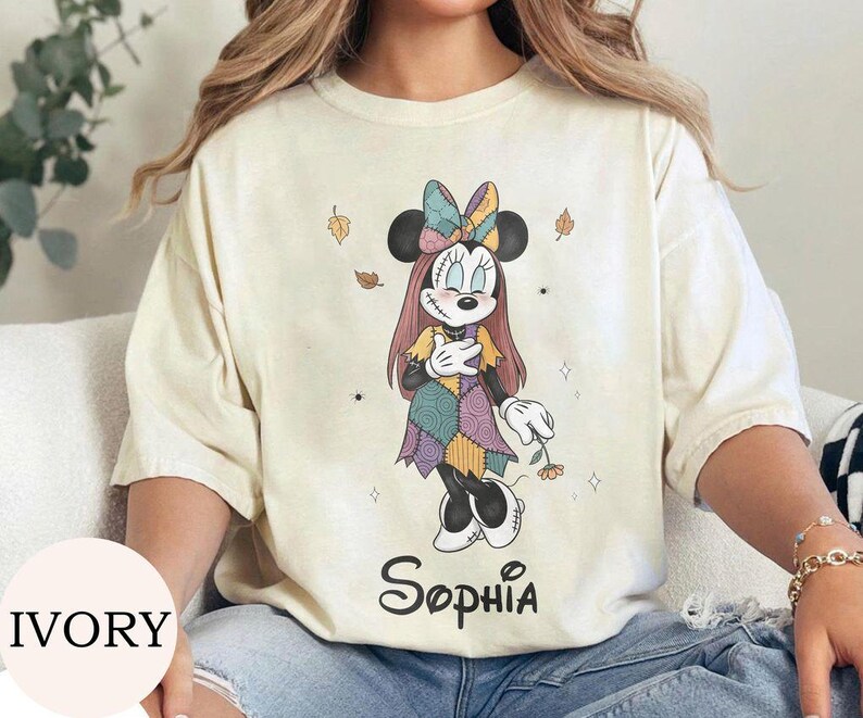 May include: An ivory t-shirt featuring a graphic of a character resembling Minnie Mouse dressed in a patchwork-style dress and bow, with long hair and stitched details. The name Sophia is printed in black script below the character illustration.