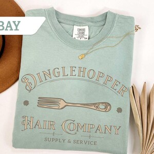 May include: A pale green t-shirt featuring the words "Dinglehopper Hair Company" and a fork illustration. The shirt also has the text "Supply & Service". A gold necklace and a brown hat are also in the image.