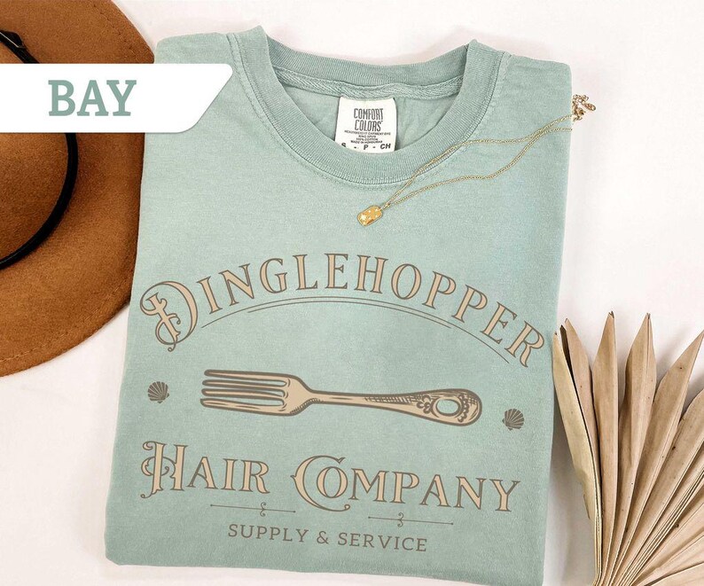 May include: A pale green t-shirt featuring the words "Dinglehopper Hair Company" and a fork illustration. The shirt also has the text "Supply & Service". A gold necklace and a brown hat are also in the image.