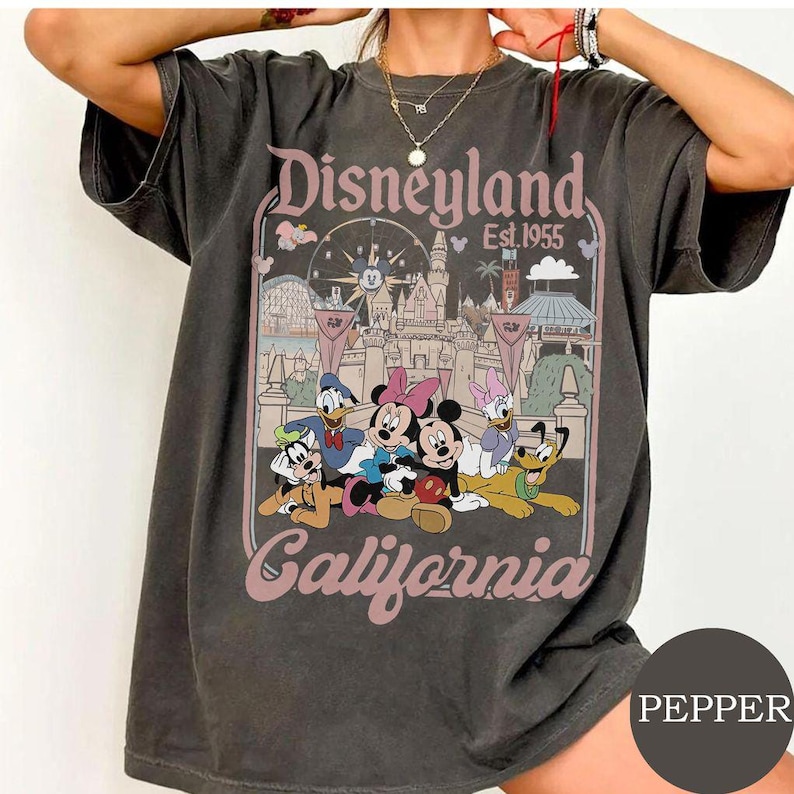 May include: A charcoal gray short-sleeve t-shirt featuring a graphic print of Disneyland California with the text Disneyland Est. 1955 California. The design includes illustrations of Mickey Mouse, Minnie Mouse, Donald Duck, Daisy Duck, Goofy, and Pluto.