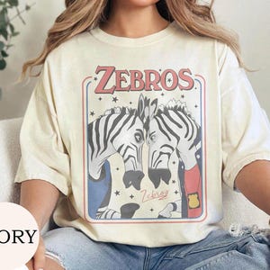 May include: An ivory t-shirt featuring a graphic print of two zebras facing each other inside a rectangular frame. The text ZEBROS appears in red at the top, with smaller cursive text below the zebras. The zebras wear blue and red clothing.