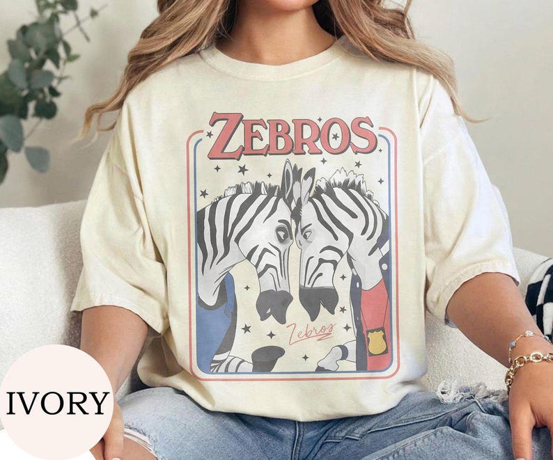 May include: An ivory t-shirt featuring a graphic print of two zebras facing each other inside a rectangular frame. The text ZEBROS appears in red at the top, with smaller cursive text below the zebras. The zebras wear blue and red clothing.