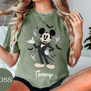 May include: A moss green t-shirt featuring a graphic of Mickey Mouse dressed in a black and white pinstriped suit, surrounded by small ghosts, bats, and stars. The name Tommy is printed in white cursive script below the central character graphic.