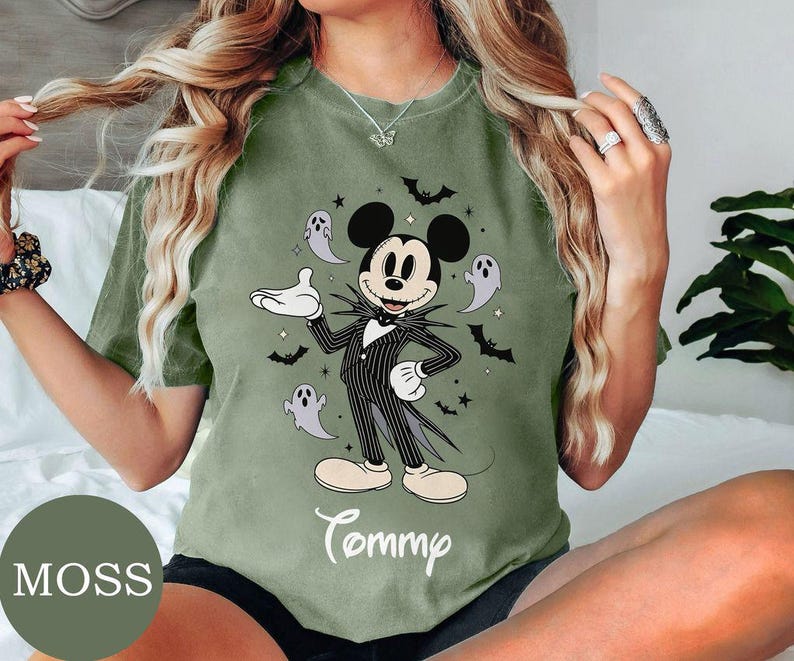 May include: A moss green t-shirt featuring a graphic of Mickey Mouse dressed in a black and white pinstriped suit, surrounded by small ghosts, bats, and stars. The name Tommy is printed in white cursive script below the central character graphic.