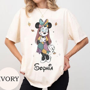 May include: An ivory short-sleeve t-shirt featuring a graphic of a cartoon mouse character with stitched facial details, wearing a patchwork dress and bow, holding a flower. The name Sophia is printed in black script below the character.