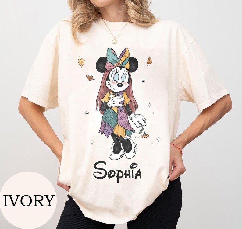 May include: An ivory short-sleeve t-shirt featuring a graphic of a cartoon mouse character with stitched facial details, wearing a patchwork dress and bow, holding a flower. The name Sophia is printed in black script below the character.