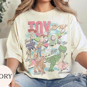 May include: An ivory short-sleeve t-shirt featuring a colorful graphic print of Toy Story characters including Woody, Buzz Lightyear, Jessie, Rex, Hamm, Bullseye, and Slinky Dog. The text reads Toy Story, Jessie Since 1995, Woody, Buzz, Rex, Hamm, and Bullseye.