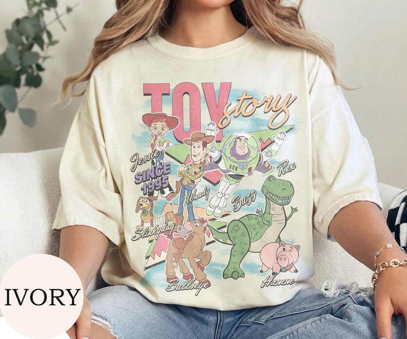 May include: An ivory short-sleeve t-shirt featuring a colorful graphic print of Toy Story characters including Woody, Buzz Lightyear, Jessie, Rex, Hamm, Bullseye, and Slinky Dog. The text reads Toy Story, Jessie Since 1995, Woody, Buzz, Rex, Hamm, and Bullseye.