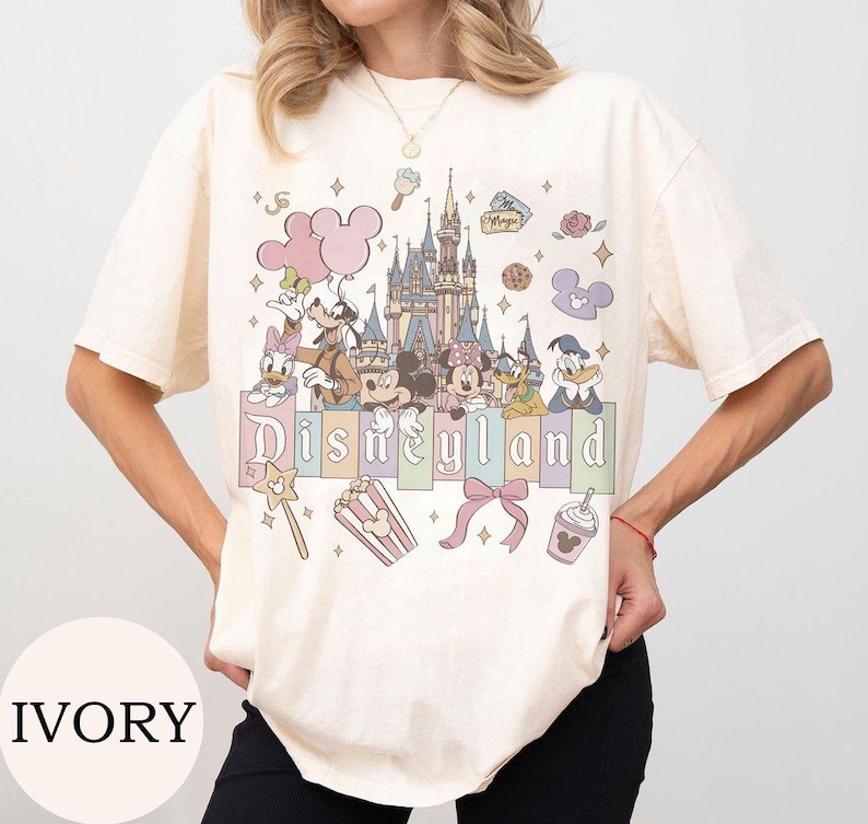 May include: An ivory t-shirt featuring a graphic print of a castle, Mickey Mouse, Minnie Mouse, Donald Duck, Daisy Duck, Goofy, and Pluto. The design includes the text Disneyland, pink balloons, a popcorn bucket, a bow, and a drink cup.