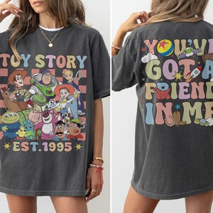 May include: Dark gray t-shirt featuring a colorful graphic of Toy Story characters with the text "TOY STORY EST. 1995" on the front. The back of the shirt has the text "YOU'VE GOT A FRIEND IN ME" with various illustrations.