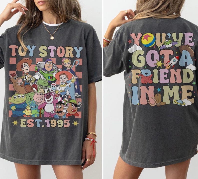 May include: Dark gray t-shirt featuring a colorful graphic of Toy Story characters with the text "TOY STORY EST. 1995" on the front. The back of the shirt has the text "YOU'VE GOT A FRIEND IN ME" with various illustrations.