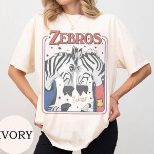 May include: An ivory short-sleeve t-shirt featuring a graphic print of two zebras facing each other. The text "ZEBROS" appears at the top in a retro font, with a smaller "Zebros" signature below. The zebras wear clothing and a police badge.