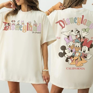 Puede incluir: Camiseta color crema con la palabra "Disneyland" en letras coloridas en la parte delantera, con personajes de Disney. La parte trasera de la camiseta muestra m&aacute;s personajes y las palabras "Disneyland California Est 1955."