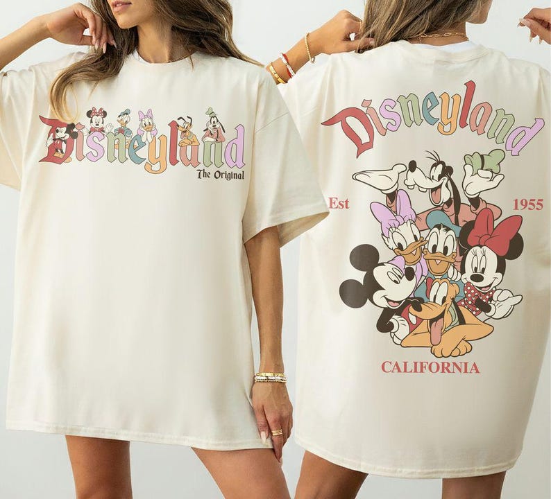 Puede incluir: Camiseta color crema con la palabra "Disneyland" en letras coloridas en la parte delantera, con personajes de Disney. La parte trasera de la camiseta muestra m&aacute;s personajes y las palabras "Disneyland California Est 1955."