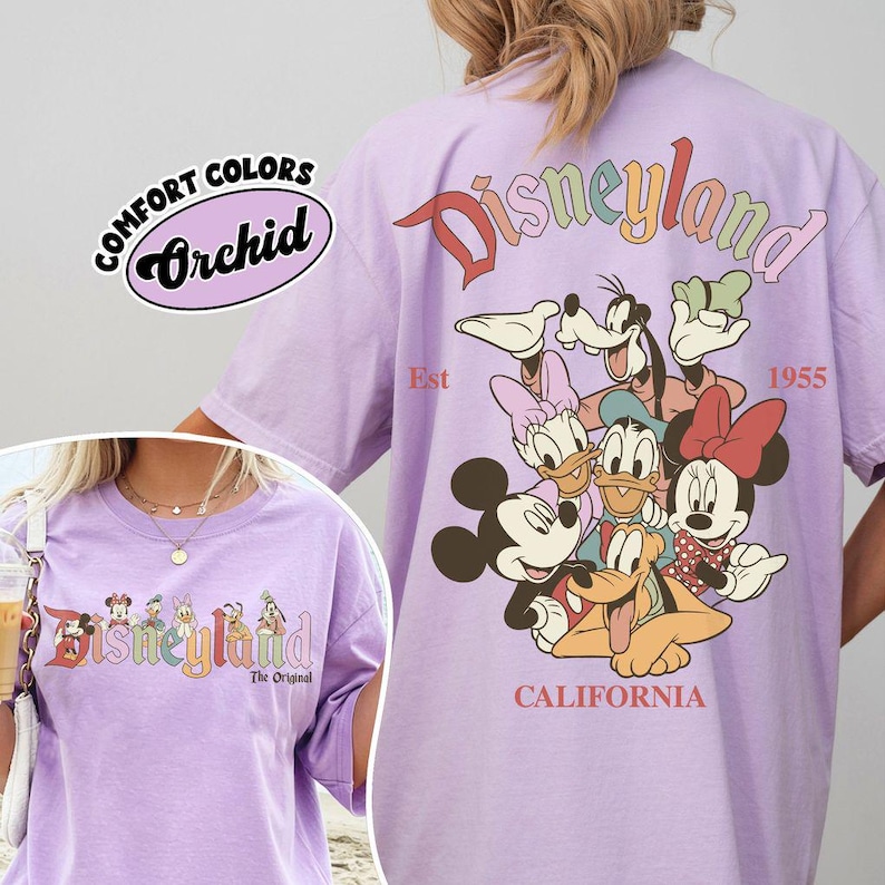 Puede incluir: Una camiseta de color lila claro con la palabra "Disneyland" en colores arco&iacute;ris y un gr&aacute;fico de personajes de Disney. La camiseta tambi&eacute;n presenta el texto "Est 1955" y "California". La camiseta est&aacute; etiquetada como "Comfort Colors Orchid".
