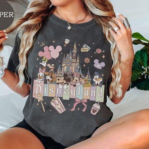 May include: A charcoal gray t-shirt featuring a graphic print of a castle, Mickey Mouse, Minnie Mouse, Donald Duck, Daisy Duck, Goofy, and Pluto. The design includes the text Disneyland, popcorn, a bow, and balloons in pastel colors.