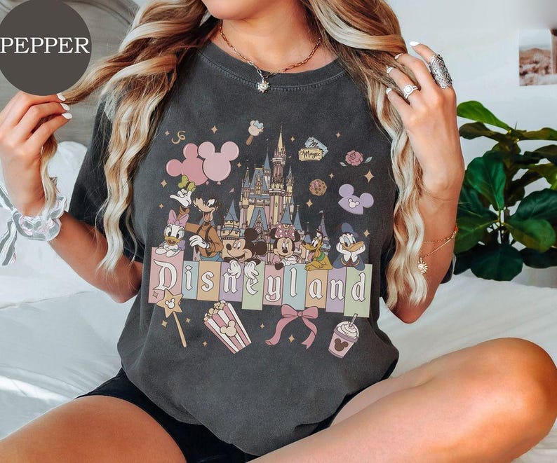 May include: A charcoal gray t-shirt featuring a graphic print of a castle, Mickey Mouse, Minnie Mouse, Donald Duck, Daisy Duck, Goofy, and Pluto. The design includes the text Disneyland, popcorn, a bow, and balloons in pastel colors.