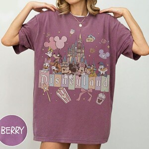 May include: A berry-colored short-sleeve t-shirt featuring a graphic print of Disneyland with a castle, Mickey Mouse, Minnie Mouse, Donald Duck, Daisy Duck, Goofy, and Pluto. The design includes icons like balloons, popcorn, and a bow with the text Disneyland.