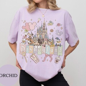May include: A light purple short-sleeve t-shirt featuring a graphic print of a castle, Mickey Mouse, Minnie Mouse, Goofy, Donald Duck, Daisy Duck, and Pluto. The text Disneyland is printed across the center with icons of balloons, popcorn, and a bow.
