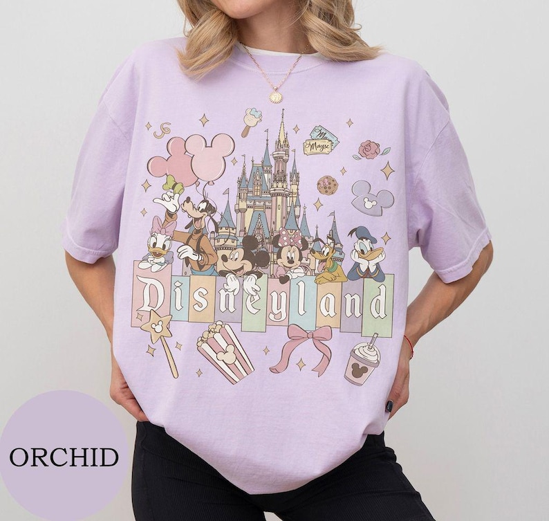 May include: A light purple short-sleeve t-shirt featuring a graphic print of a castle, Mickey Mouse, Minnie Mouse, Goofy, Donald Duck, Daisy Duck, and Pluto. The text Disneyland is printed across the center with icons of balloons, popcorn, and a bow.