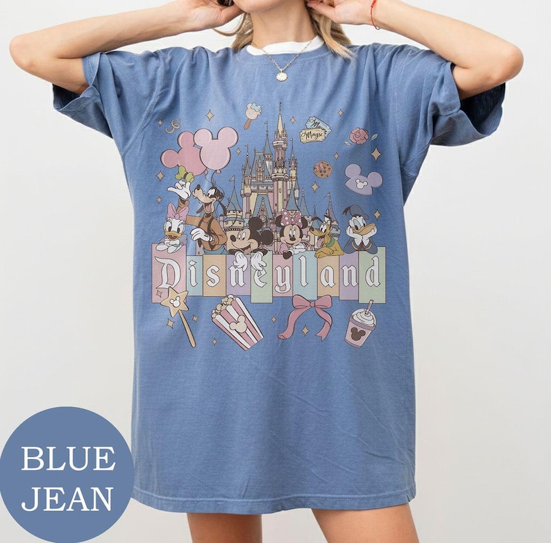 May include: A blue jean colored t-shirt featuring a graphic print of the Disneyland castle and various cartoon characters including Mickey, Minnie, Goofy, Donald, and Daisy. The design includes pastel colored accents, a bow, and snacks.