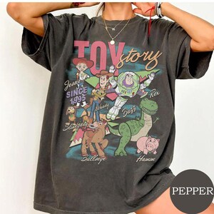 May include: A charcoal gray short-sleeve t-shirt featuring a graphic print of Toy Story characters including Woody, Buzz Lightyear, Jessie, Rex, Hamm, Slinky Dog, and Bullseye. The text reads Toy Story, Jessie Since 1995, Woody, Buzz, Rex, Hamm, and Bullseye.