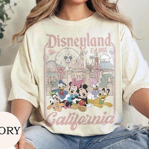 May include: An ivory short-sleeve t-shirt featuring a graphic print of Disneyland California Est. 1955. The design includes Mickey Mouse, Minnie Mouse, Donald Duck, Daisy Duck, Goofy, and Pluto in front of the castle and a Ferris wheel.