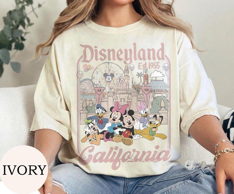 May include: An ivory short-sleeve t-shirt featuring a graphic print of Disneyland California Est. 1955. The design includes Mickey Mouse, Minnie Mouse, Donald Duck, Daisy Duck, Goofy, and Pluto in front of the castle and a Ferris wheel.