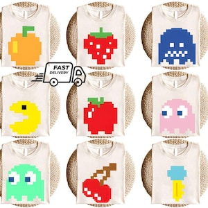 以下が含まれることがあります： ピクセル化された果物とビデオゲームのキャラクターのデザインが施された、9枚のオフホワイトのTシャツ。デザインには、オレンジ、イチゴ、青い幽霊、黄色のパックマン、赤いリンゴ、ピンクの幽霊、さくらんぼ、アイスクリームコーンが含まれています。「FAST DELIVERY」の文字も表示されています。