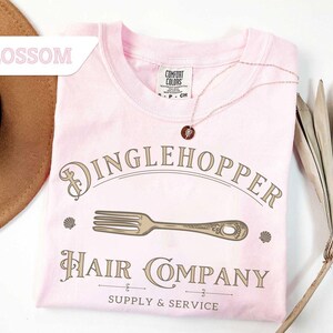 May include: A pale pink Comfort Colors t-shirt featuring the words "Dinglehopper Hair Company" in a vintage font. The design includes a fork graphic and the words "Supply & Service". A gold necklace with a pendant is draped on the shirt.