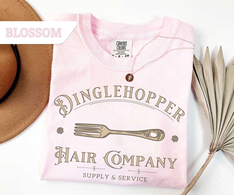 May include: A pale pink Comfort Colors t-shirt featuring the words "Dinglehopper Hair Company" in a vintage font. The design includes a fork graphic and the words "Supply & Service". A gold necklace with a pendant is draped on the shirt.