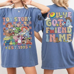 May include: Blue t-shirt featuring a colorful graphic of Toy Story characters with the text "TOY STORY EST. 1995" on the front. The back of the shirt has the phrase "YOU'VE GOT A FRIEND IN ME" in a retro font.