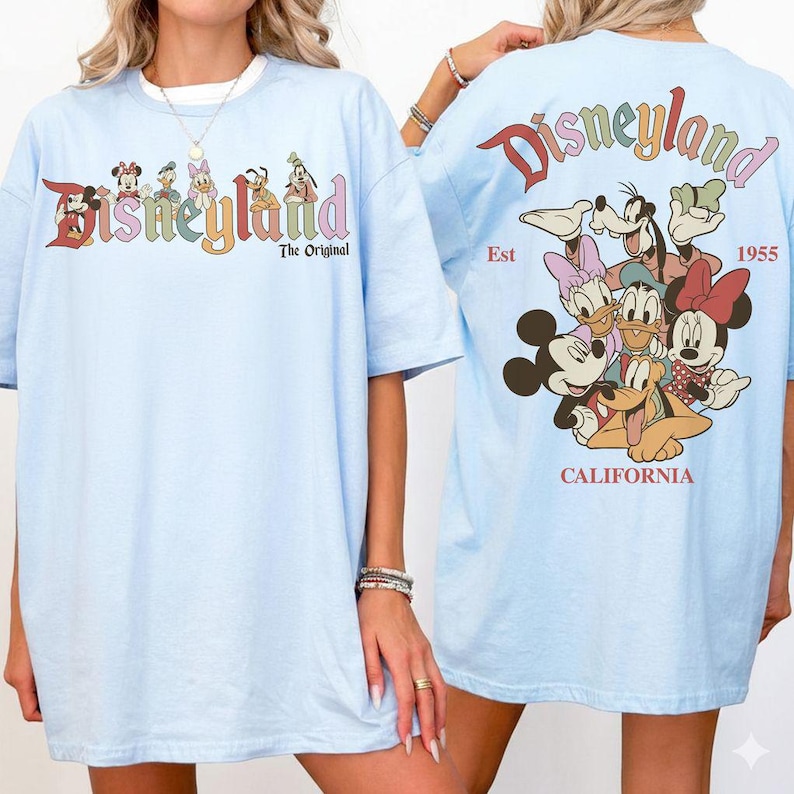 Puede incluir: Camisetas de color azul claro con un dise&ntilde;o de Disneyland. La parte delantera de la camiseta tiene la palabra "Disneyland" en letras coloridas con personajes de Disney. La parte trasera tiene la palabra "Disneyland" en un dise&ntilde;o curvo con personajes de Disney y el texto "Est 1955 California".