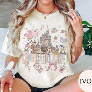 May include: An ivory t-shirt featuring a graphic print of a castle, Mickey Mouse, Minnie Mouse, Donald Duck, Daisy Duck, Goofy, and Pluto. The design includes pastel-colored balloons, a popcorn bucket, a bow, and the text Disneyland.