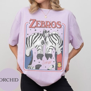 May include: A light purple short-sleeve t-shirt featuring a graphic print of two zebras facing each other. The text ZEBROS is printed in red above the zebras, with small stars surrounding them. One zebra wears a blue vest and the other a red shirt.