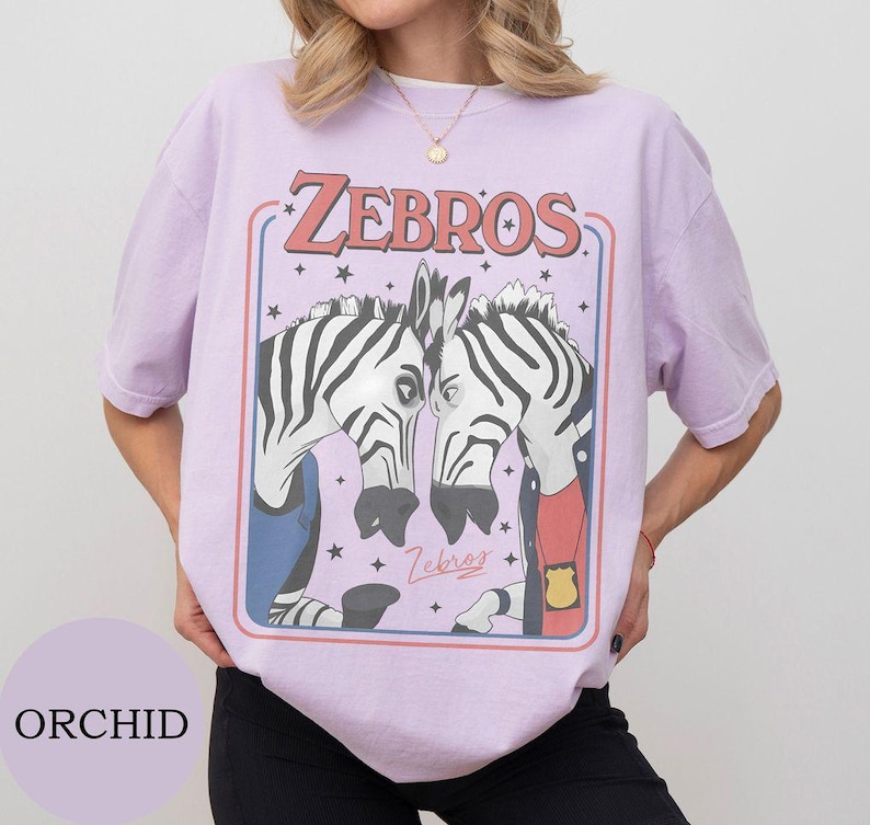 May include: A light purple short-sleeve t-shirt featuring a graphic print of two zebras facing each other. The text ZEBROS is printed in red above the zebras, with small stars surrounding them. One zebra wears a blue vest and the other a red shirt.