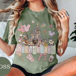 May include: A moss green t-shirt featuring a graphic print of a castle, Mickey Mouse, Minnie Mouse, Donald Duck, Daisy Duck, Goofy, and Pluto. The text "Disneyland" is printed across the center, surrounded by icons of balloons, popcorn, and a bow.