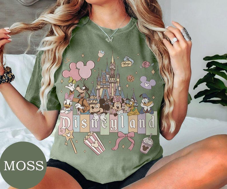 May include: A moss green t-shirt featuring a graphic print of a castle, Mickey Mouse, Minnie Mouse, Donald Duck, Daisy Duck, Goofy, and Pluto. The text "Disneyland" is printed across the center, surrounded by icons of balloons, popcorn, and a bow.