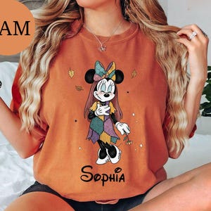 May include: An orange crewneck t-shirt featuring a graphic of a cartoon mouse character with stitched facial details, wearing a patchwork dress and bow, holding a small flower. The name Sophia is printed in black script below the character.