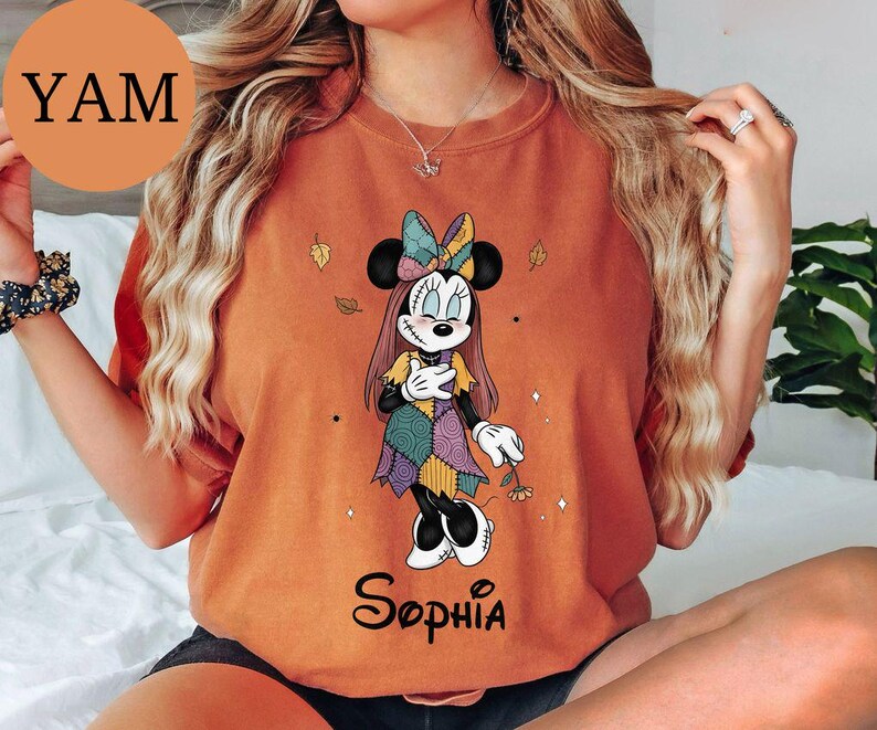May include: An orange crewneck t-shirt featuring a graphic of a cartoon mouse character with stitched facial details, wearing a patchwork dress and bow, holding a small flower. The name Sophia is printed in black script below the character.