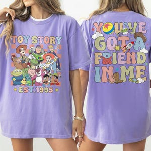 May include: A lilac-colored t-shirt featuring a colorful graphic of Toy Story characters on the front. The back of the shirt displays the phrase "You've Got A Friend In Me" in a retro font. The shirt also has the text "EST.1995".