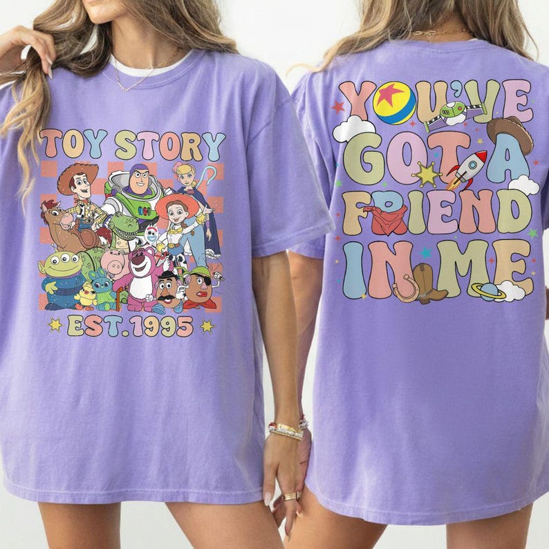 May include: A lilac-colored t-shirt featuring a colorful graphic of Toy Story characters on the front. The back of the shirt displays the phrase "You've Got A Friend In Me" in a retro font. The shirt also has the text "EST.1995".