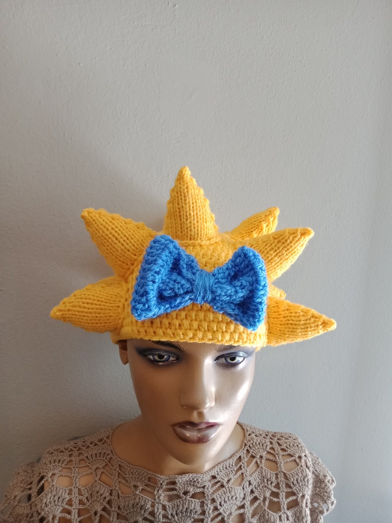 May include: A yellow knitted hat shaped like a sun with a blue knitted bow on top.