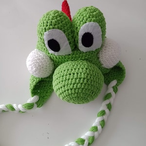 Puede incluir: Sombrero de crochet verde con trenzas de hilo blanco y verde, con una cara de dinosaurio de dibujos animados con ojos blancos y una punta roja en la parte superior.