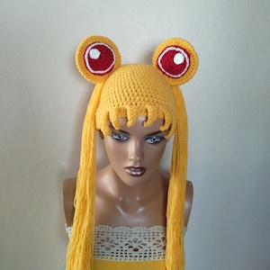 Può includere: Un cappello di uncinetto giallo con lunghe code di filo giallo e orecchie di uncinetto rosse e bianche. Il cappello è su una testa di manichino.