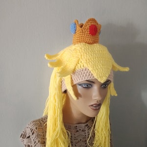 May include: A yellow crocheted hat with a crown on top. The crown is brown with red and blue accents. The hat has long yellow yarn strands hanging down the back.