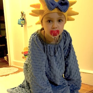 May include: A child wearing a blue fuzzy pajama top and a yellow crocheted sun hat with a blue bow. The child is wearing a red pacifier.