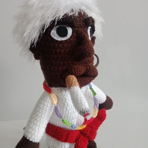 Puede incluir: Una muñeca de crochet con pelo blanco, piel marrón y un atuendo blanco. La muñeca lleva una faja roja y un collar de cuentas de colores. La muñeca sostiene un cigarrillo en la boca.