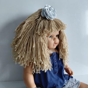 May include: A doll with a handmade wig made of long, beige yarn strands and a silver fabric flower. The doll is wearing a blue dress with button details and a white floral embroidered hemline. The doll has blue eyes.