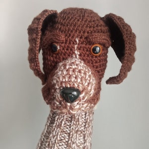 May include: A brown and white crocheted dog puppet with brown eyes and a black nose. The puppet has a long brown and white crocheted neck.
